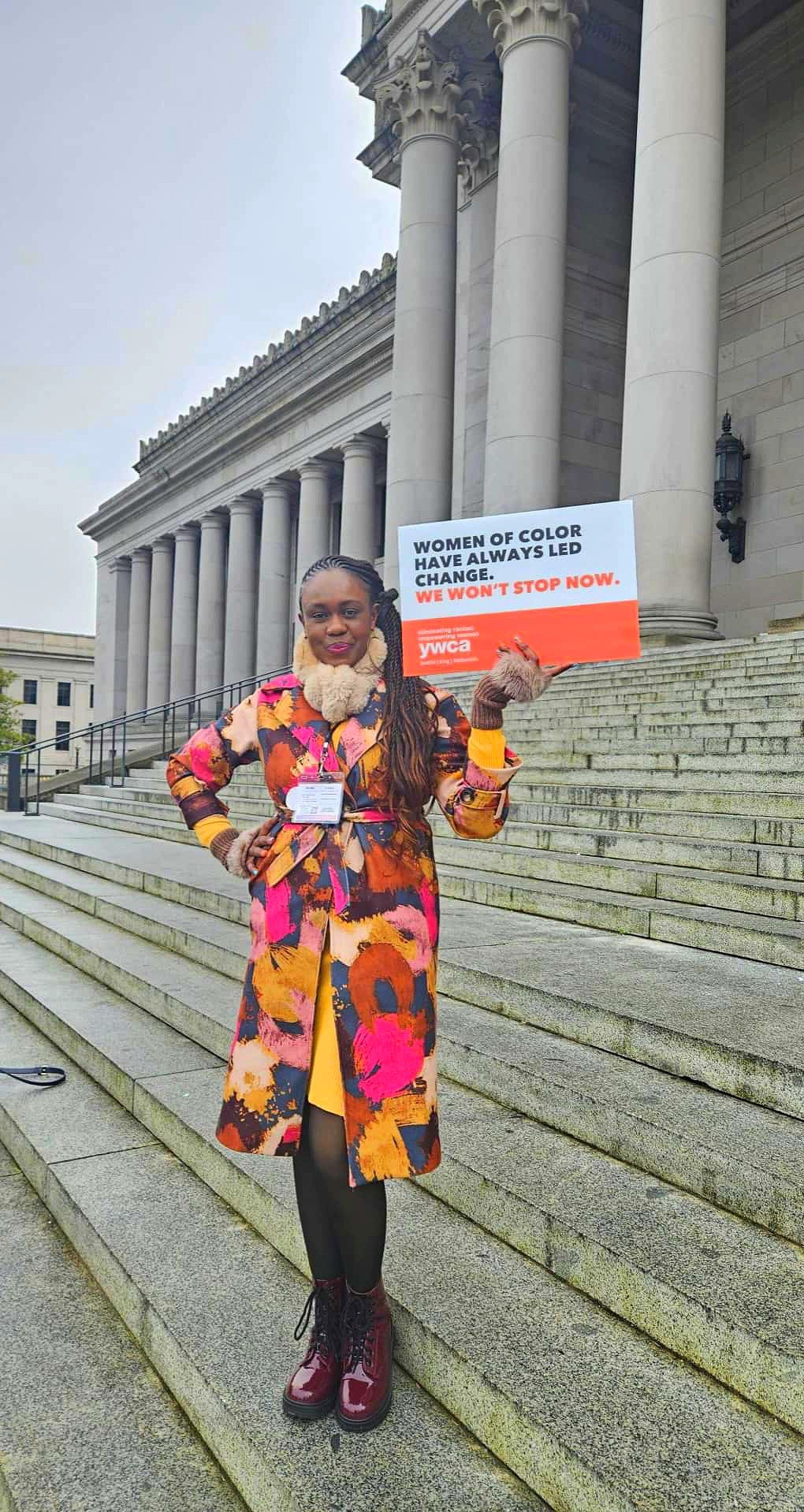 Advocate standing on the steps of a civic building holding a sign calling for women of color to continue leading change.